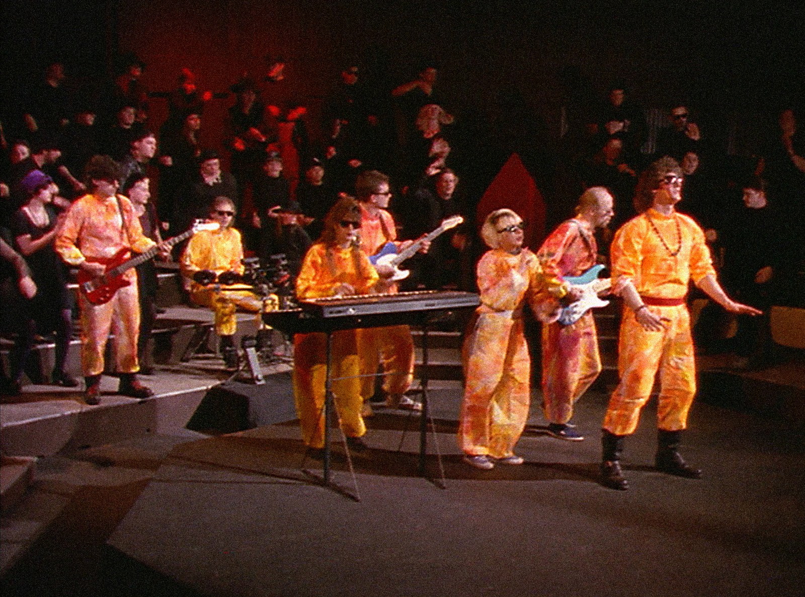 The Jupiter Sheep on a stepped stage in orange suits, the crowd behind them dressed in black